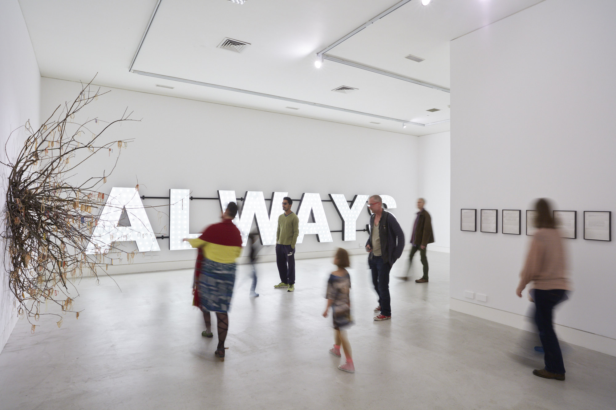 A white-walled gallery with people blurred in motion viewing artworks. On the far wall is a large illuminated letter that reads ‘ALWAYS’. On the left wall is a thick cluster of branches coming out of the wall like a root network with elastic bands hung over the branches.