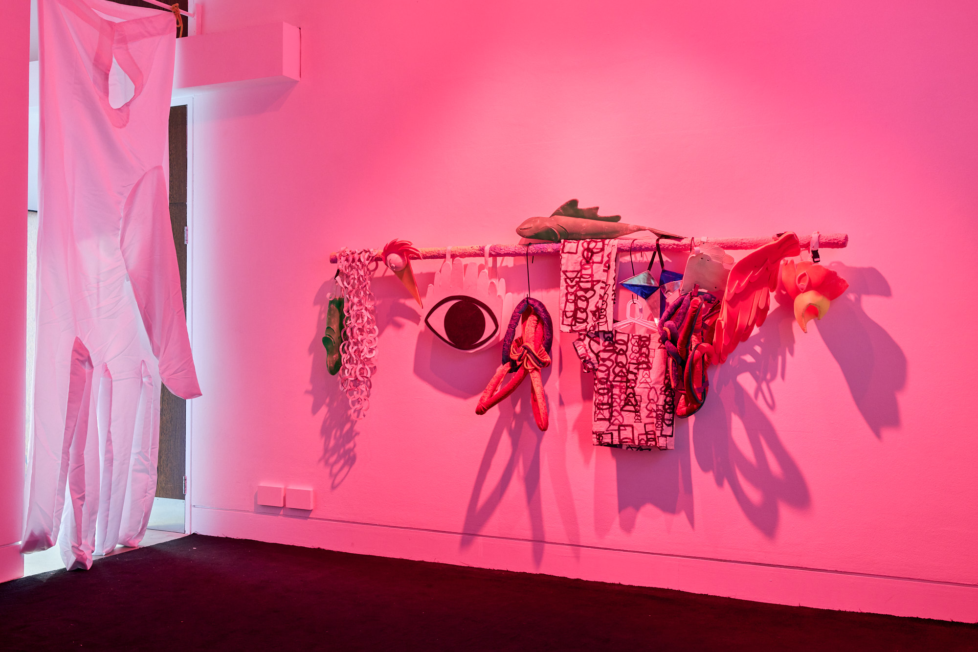 A set of sculptural costumes hanging in a line, a pink light eliminates the gallery space.