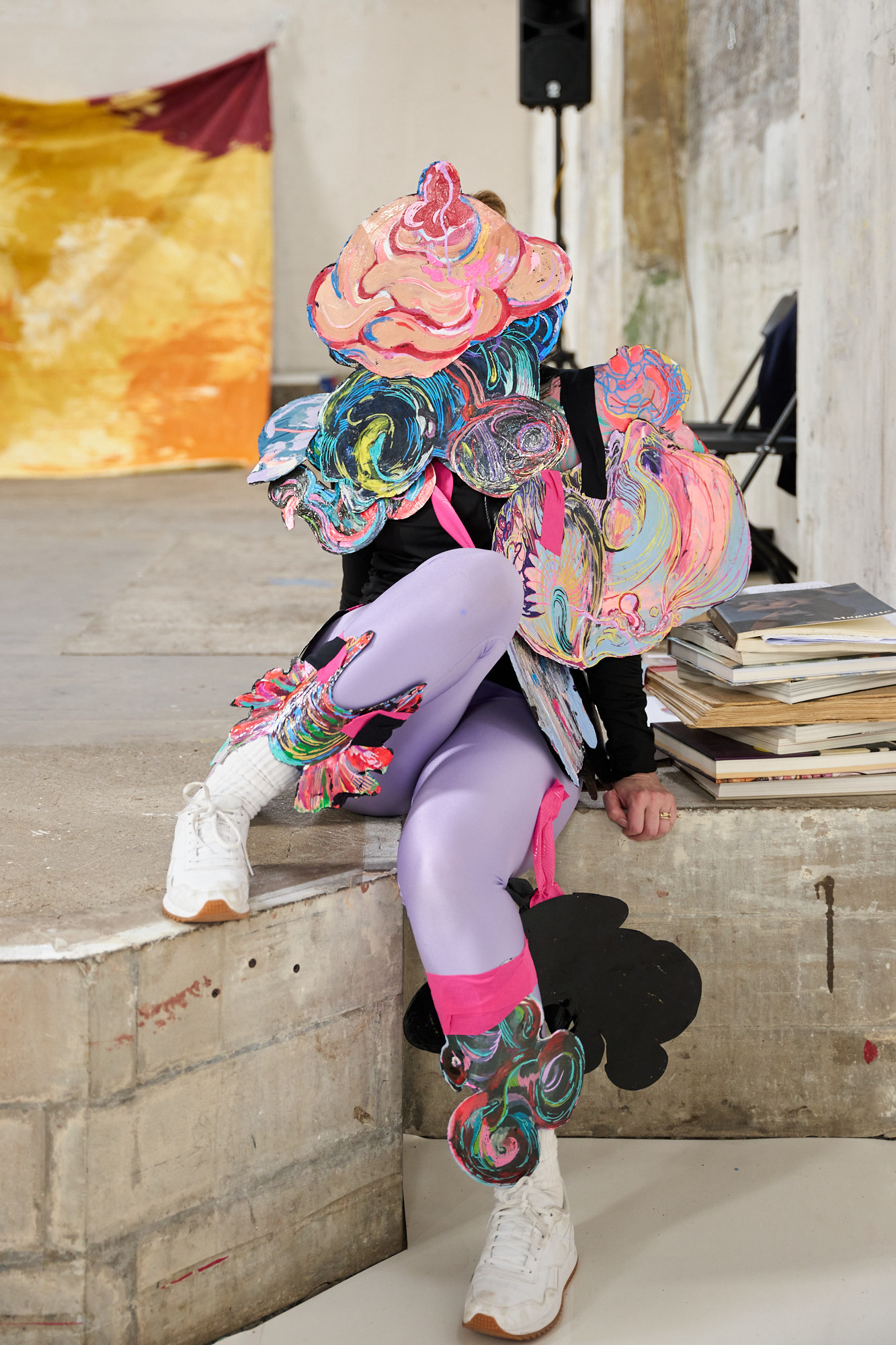 A performer in lilac leggings with colourful painted accessories covering their face sits with one leg resting above the other.