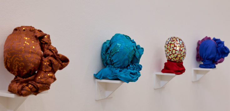 Close up image of a four small bright artworks mounted on a white wall. Each is a twisted fabric sculpture on a small white wall shelf in different colours, from left to right: Burnt orange; turquoise; red with gold sequins; and royal blue and mauve.