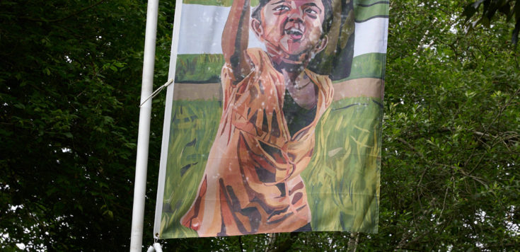 In a lush green park, a white flagpole depicts a colourful flag of an oil painting of a reaching child.