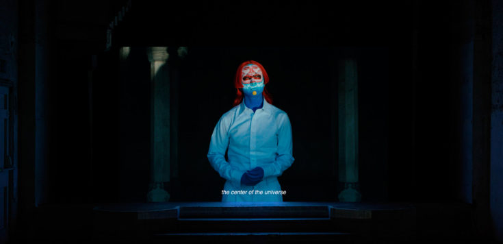 A projected film in a dark space displays a character dressed in white with a blue and red painted face, red hair and subtitles.