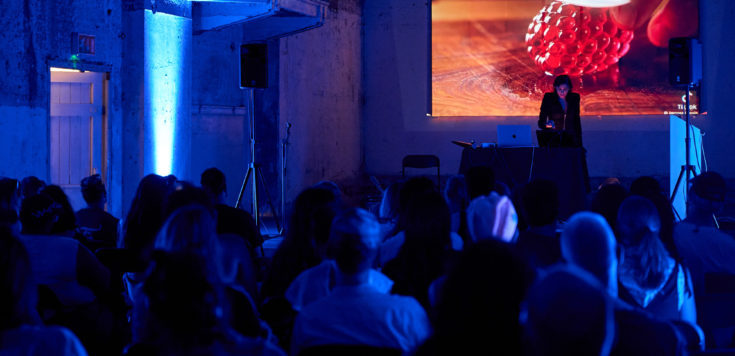 A Zein Majali operates audio equipment behind a laptop on a dimly lit stage with a AI visuals of a glass raspberry being sliced projected on the screen behind them.