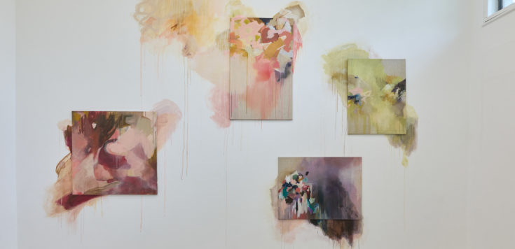 A gallery wall displaying abstract artworks in soft colors, with larger pieces draping in vibrant drips and smaller framed pieces to the left.