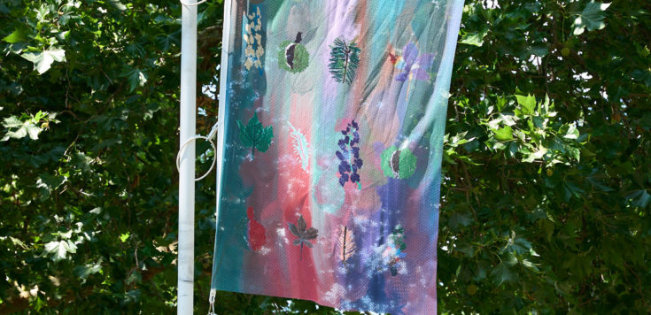 A colourful flag with abstract leaf and floral patterns hangs on a white pole, set in a sunny park with green trees.